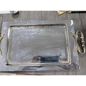 May include: A rectangular silver-toned metal serving tray featuring a polished surface with visible signs of wear and oxidation. The tray includes two ornate, gold-toned metal handles attached to the sides, each with a decorative scroll design.