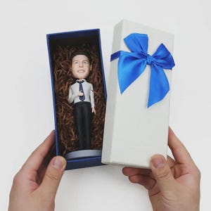 May include: A bobblehead figurine of a person in a white shirt and black trousers, with a blue tie, inside a blue box with brown packing material. A white gift box with a blue ribbon is next to it.