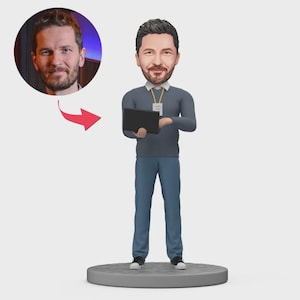 May include: A custom bobblehead figurine of a man holding a tablet, wearing a gray sweater, blue pants, and sneakers. A photo of the man is in a circle above the figurine, connected by a red arrow. The figurine stands on a gray circular base.