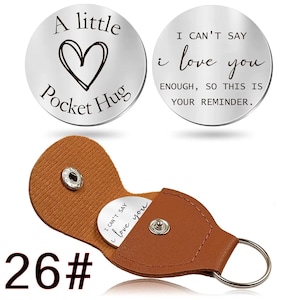 Puede incluir: Dos fichas de bolsillo plateadas con mensajes grabados. Una dice "A little Pocket Hug" con un corazón, la otra dice "I can't say I love you enough, so this is your reminder." Las fichas están en un estuche de llavero de cuero marrón.