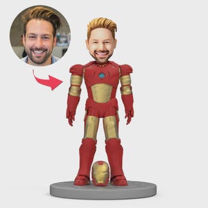 May include: A custom bobblehead figurine of a man in an Iron Man suit, with a detached helmet. The red and gold suit is detailed, and the head is a likeness of the person. The figurine stands on a grey circular base.