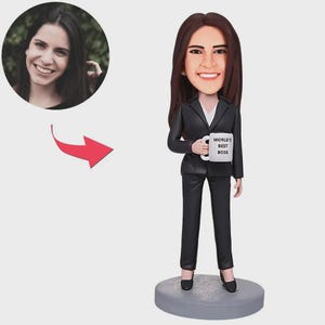 May include: A figurine of a woman in a black suit holding a mug that says "WORLD'S BEST BOSS." The figurine has a smiling face and long brown hair. A photo of a smiling woman is in a circle in the upper left corner.