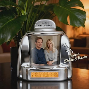 Op de afbeelding: Een zilverkleurige, op maat gemaakte mini-jukebox met een foto van een stel erin. De jukebox heeft een selectiepaneel met letters en cijfers. De tekst "Custom Photo & Music" staat op de zijkant.