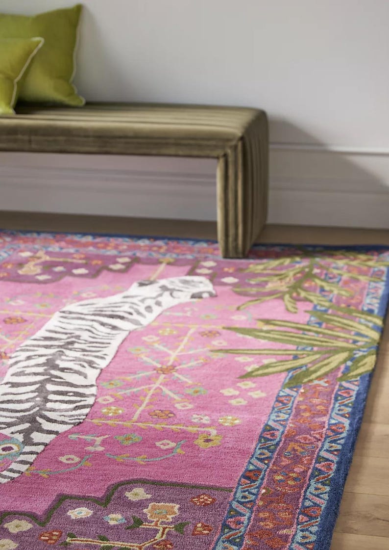 May include: A vibrant pink rug with a zebra stripe design and botanical accents. The rug features a blue border and a decorative pattern. A green bench and pillows are in the background.