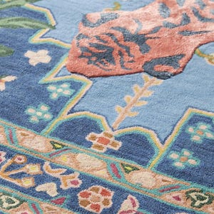 May include: Close-up of a blue area rug with a floral design. The rug features a light blue center with a large coral-colored floral motif. The border has a detailed pattern with various colors, including pink, green, and yellow.