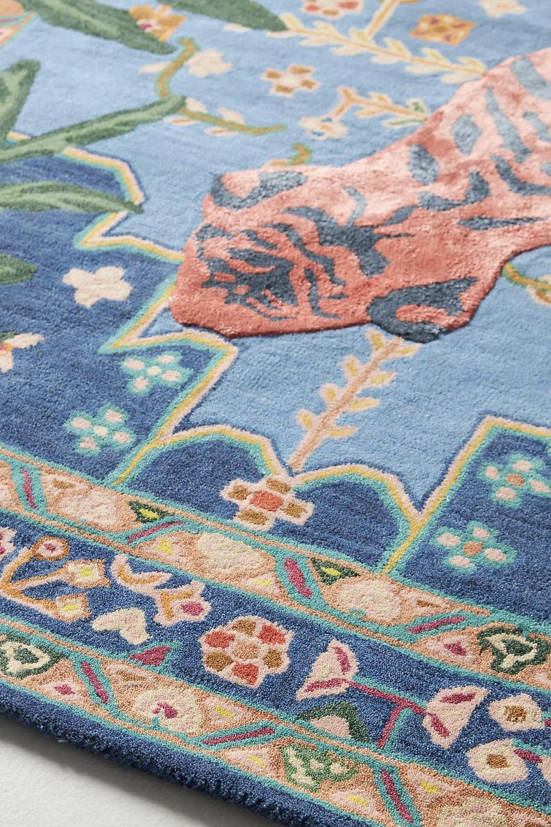 May include: Close-up of a blue area rug with a floral design. The rug features a light blue center with a large coral-colored floral motif. The border has a detailed pattern with various colors, including pink, green, and yellow.