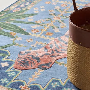 May include: A light blue rug with a floral design and a woven basket with a brown leather trim. The rug features a pattern of flowers, leaves, and geometric shapes in shades of blue, pink, and green.