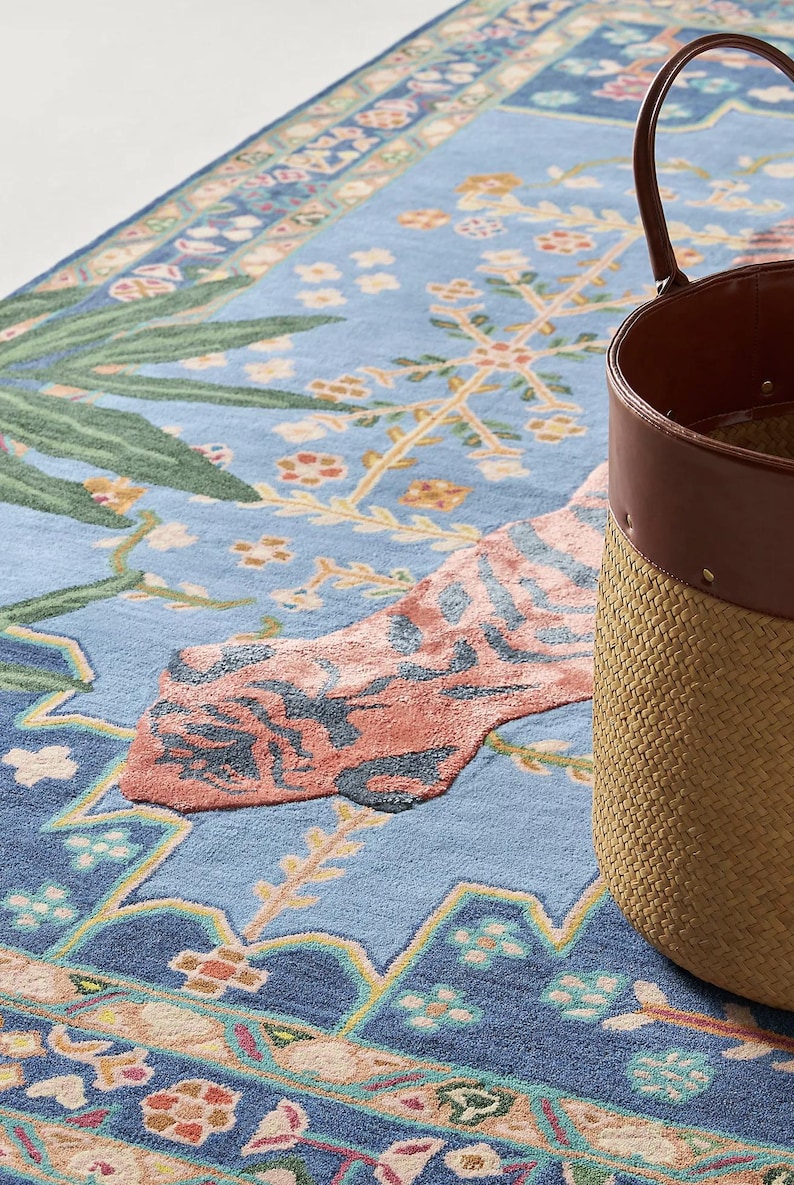 May include: A light blue rug with a floral design and a woven basket with a brown leather trim. The rug features a pattern of flowers, leaves, and geometric shapes in shades of blue, pink, and green.