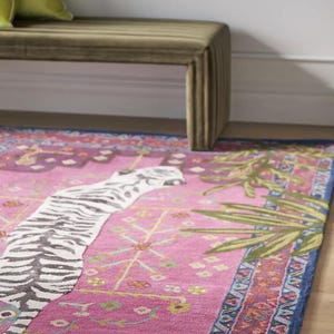 May include: A vibrant pink area rug with a zebra stripe design and a blue border. The rug features a detailed pattern with floral and geometric elements, and green palm fronds. A green bench and pillows are in the background.