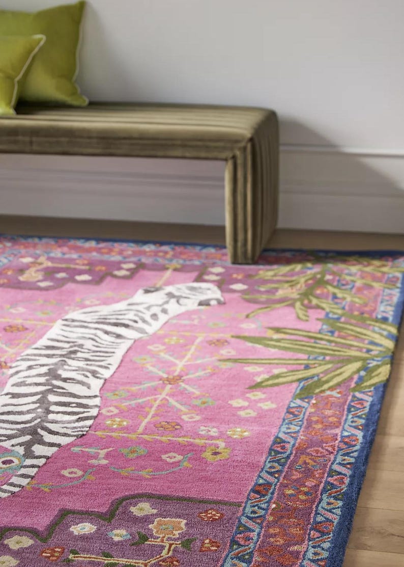 May include: A vibrant pink area rug with a zebra stripe design and a blue border. The rug features a detailed pattern with floral and geometric elements, and green palm fronds. A green bench and pillows are in the background.