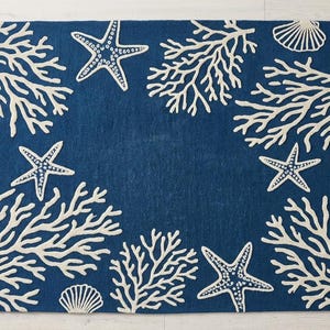May include: A rectangular area rug featuring a deep blue background with a repeating pattern of off-white coral branches, five-pointed starfish, and fan-shaped seashells arranged around the perimeter, leaving a solid blue center space.