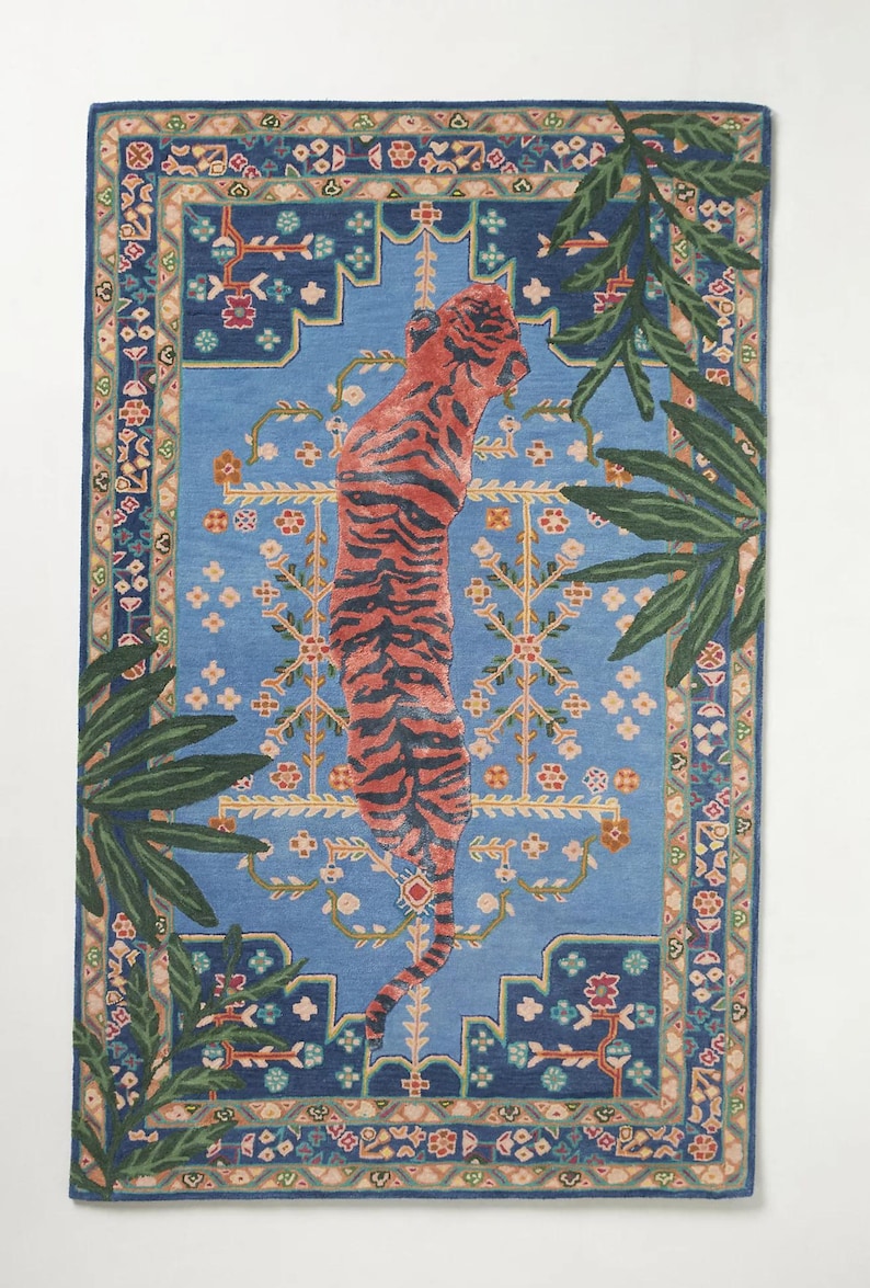 May include: Rectangular blue rug with a central design of a standing tiger with orange and black stripes. The rug has a detailed border with floral and geometric patterns, and green palm fronds frame the tiger.