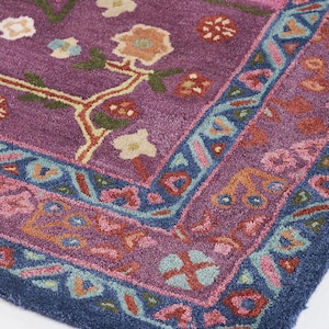 May include: Close-up of a section of a vibrantly colored rug. The rug features a deep purple central field with floral and vine motifs in shades of orange, green, and cream. A border of blue and pink surrounds the central design.