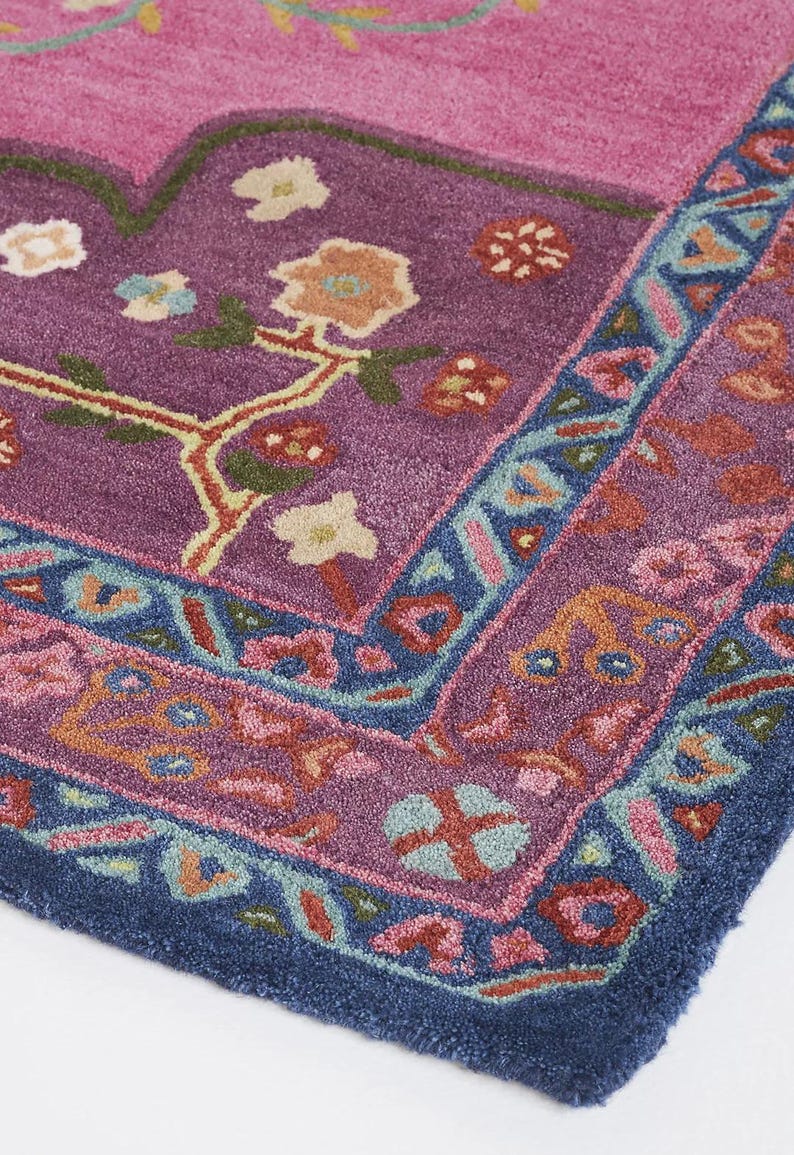 May include: Close-up of a section of a vibrantly colored rug. The rug features a deep purple central field with floral and vine motifs in shades of orange, green, and cream. A border of blue and pink surrounds the central design.