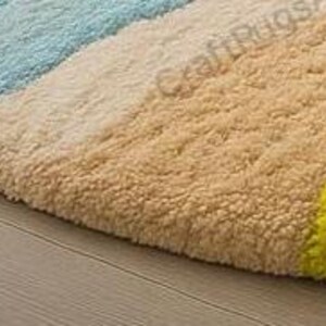 Pode incluir: Um close-up de um tapete redondo e colorido com uma superf&iacute;cie texturizada. O tapete apresenta sec&ccedil;&otilde;es azuis, bege e amarelas. O tapete est&aacute; colocado sobre um ch&atilde;o de madeira clara.