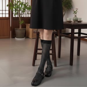 May include: Black split-toe shoes with a buckle closure are worn with knee-high socks featuring vertical stripes. The socks are paired with a black skirt. The background includes wooden furniture and a potted plant.