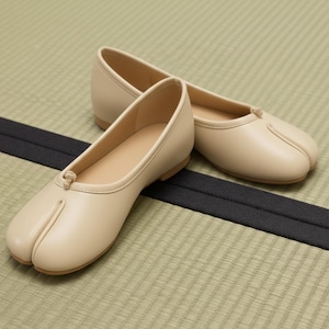 Può includere: Un paio di ballerine beige con punta divisa. Le scarpe hanno un design a punta arrotondata con una divisione per l'alluce e un piccolo dettaglio a nodo sulla parte superiore. Le ballerine sono realizzate in materiale liscio e poggiano su una suola marrone chiaro.