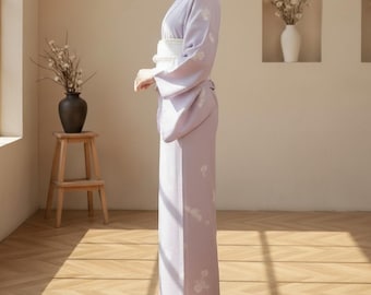 May include: A light purple kimono with white floral embroidery. The kimono is secured with a white obi belt. The sleeves are long and flowing. The garment is worn with white footwear. The background is a room with natural light.