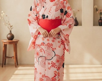 May include: A light pink Japanese kimono with a red obi belt. The kimono features a floral pattern in red and black. The sleeves are wide and the garment is ankle-length. The kimono is a traditional garment often worn during festivals.