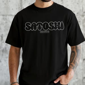 Puede incluir: Camiseta negra con la palabra "SATOSHI" en letras blancas con forma de burbujas. Debajo, la palabra "NAKAMOTO" está en un rectángulo blanco. La esquina inferior izquierda de la camiseta dice "BLACK".