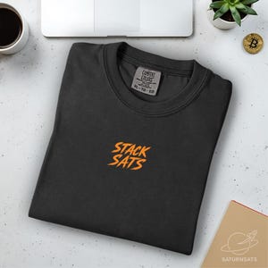 Puede incluir: Una camiseta negra de cuello redondo con el texto naranja "STACK SATS" bordado en la parte delantera. La camiseta tiene una etiqueta Comfort Colors y está doblada cuidadosamente. Una moneda de Bitcoin y una pequeña planta son visibles en el fondo.