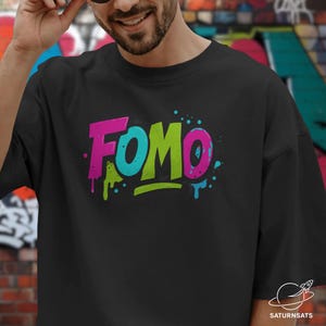 Puede incluir: Camiseta negra con la palabra "FOMO" en un diseño de estilo grafiti. La palabra "FOMO" está en rosa, turquesa y verde, con goteos y salpicaduras de pintura. La camiseta es de estilo casual y urbano.
