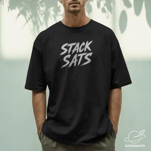 Puede incluir: Camiseta negra con el texto blanco "STACK SATS" en estilo pincelada. La camiseta tiene mangas cortas y un corte holgado. El logotipo de SaturnSats es visible en la esquina inferior derecha.