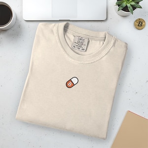 May include: Cream-colored t-shirt with a small embroidered Bitcoin pill design. The shirt is folded and has a tag that reads "Comfort Colors". A Bitcoin coin and a small plant are visible in the background.