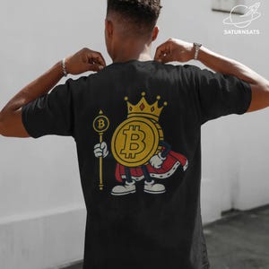 Op de afbeelding: Zwart t-shirt met een cartoon Bitcoin-personage met een gouden kroon en cape, met een scepter met een Bitcoin-symbool. Het ontwerp bevat de tekst "SATURNSATS" in de rechterbovenhoek.