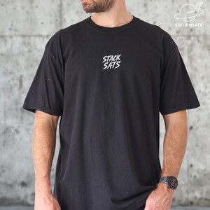 Puede incluir: Camiseta negra de manga corta con el texto blanco "STACK SATS" en el pecho. La camiseta tiene cuello redondo y un corte holgado. El logotipo de la marca "SATURNSATS" está en la esquina superior derecha.