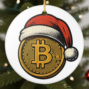 Puede incluir: Un adorno navideño redondo y blanco con un símbolo de Bitcoin dorado que lleva un gorro de Papá Noel rojo y blanco. El símbolo de Bitcoin tiene la letra "B" en el centro. Se adjunta un hilo dorado para colgar.
