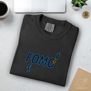 Puede incluir: Camiseta Comfort Colors gris oscuro con la palabra "FOMO" en azul y un gráfico de cohete. Una pequeña figura cuelga de la "O". La camiseta es talla XL.