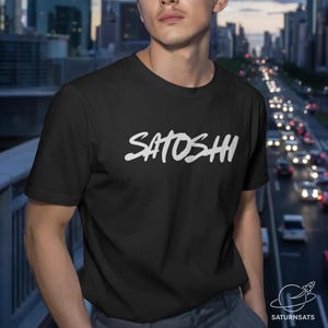 Puede incluir: Camiseta negra con la palabra "SATOSHI" en blanco, con letras estilizadas. La imagen tiene un fondo borroso de paisaje urbano. El logotipo de SaturnSats está en la esquina inferior derecha.