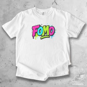 Puede incluir: Camiseta blanca con la palabra "FOMO" en un diseño estilo grafiti. El texto es en rosa brillante, azul y verde con contornos negros y goteos de pintura. La camiseta es de un material suave.