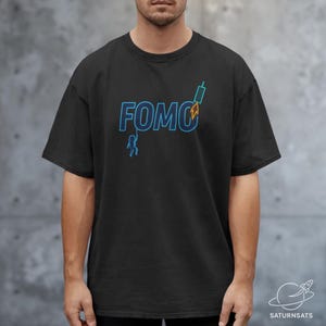 Puede incluir: Camiseta negra con un gráfico azul y verde neón. El gráfico presenta la palabra "FOMO" con una figura de palo colgando de la letra "O" y un gráfico de velas en forma de cohete. El logotipo de Saturnsats está en la esquina inferior derecha.