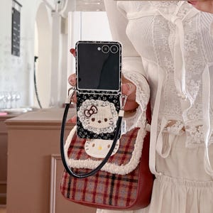 May include: A black smartphone case with a floral pattern and a Hello Kitty design is attached to a small, red and brown plaid purse. The purse has a white fluffy trim and a black strap. The phone is open.