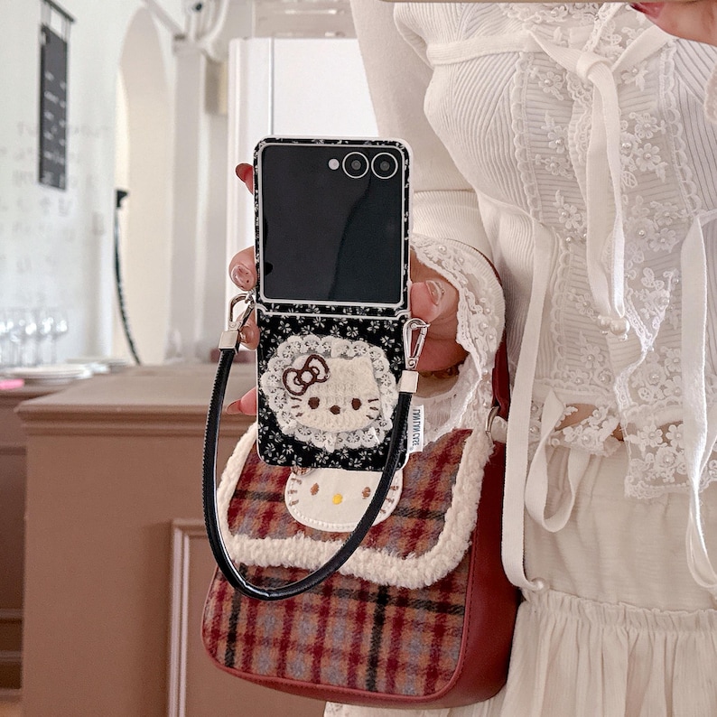 May include: A black smartphone case with a floral pattern and a Hello Kitty design is attached to a small, red and brown plaid purse. The purse has a white fluffy trim and a black strap. The phone is open.