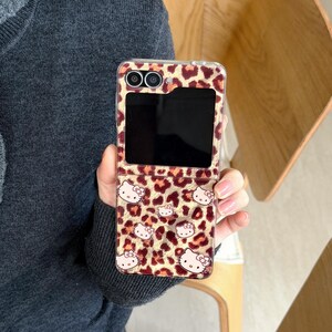 May include: A phone case featuring a leopard print pattern and Hello Kitty illustrations. The case has a tan and brown colour scheme with a clear edge. The phone case is designed to fold open and closed.