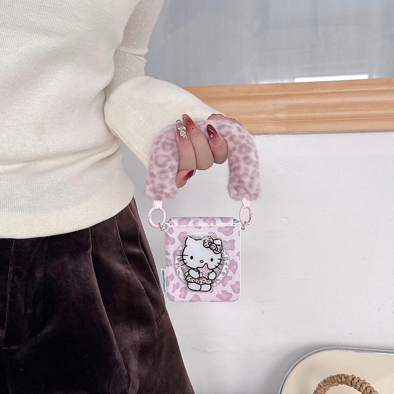 Op de afbeelding: Roze en witte AirPod-hoes met een Hello Kitty-ontwerp. De hoes heeft een roze luipaardprintpatroon en een pluizige roze handgreep. De hoes wordt vastgehouden door een persoon die een witte trui en een bruine broek draagt.