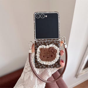 May include: Brown floral patterned phone case with a brown Hello Kitty applique and a white lace trim. The case has a brown leather strap and silver hardware. The phone screen is black.
