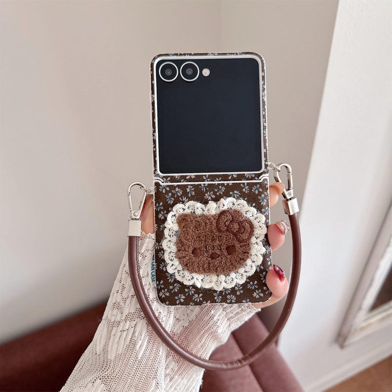 May include: Brown floral patterned phone case with a brown Hello Kitty applique and a white lace trim. The case has a brown leather strap and silver hardware. The phone screen is black.
