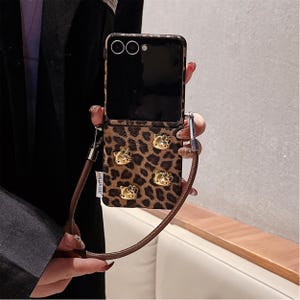 May include: A brown and black leopard print phone case with gold-colored Hello Kitty embellishments. The case has a matching brown strap and the text "LAN LAN CASE". The phone is open, revealing the screen.