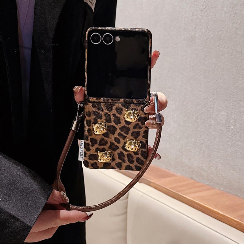 May include: A brown and black leopard print phone case with gold-colored Hello Kitty embellishments. The case has a matching brown strap and the text "LAN LAN CASE". The phone is open, revealing the screen.