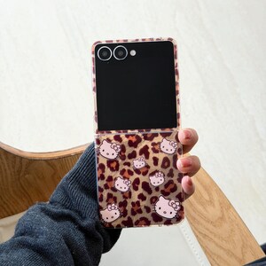 May include: A clear phone case featuring a leopard print pattern and pink Hello Kitty illustrations. The phone case is open, displaying a black screen. The case is held in a hand.