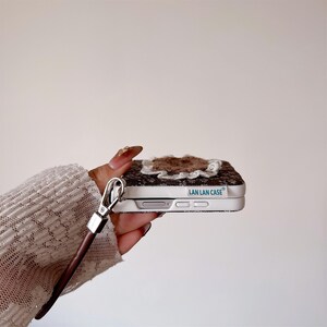 May include: A patterned phone case in brown and white, featuring a floral embellishment. It has a silver clasp and a brown leather strap. The text "LAN LAN CASE" is printed on the side of the case.