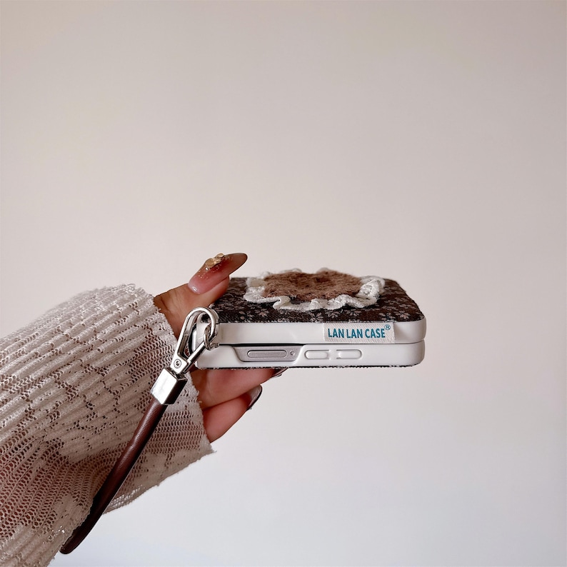 May include: A patterned phone case in brown and white, featuring a floral embellishment. It has a silver clasp and a brown leather strap. The text "LAN LAN CASE" is printed on the side of the case.