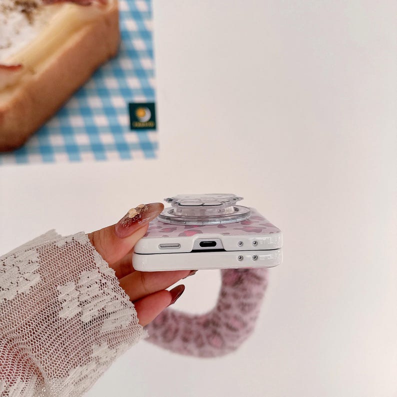 Op de afbeelding: Een witte telefoonhoes met een roze luipaardprint en een doorzichtige popsocket. De telefoonhoes wordt vastgehouden door een persoon met een witte kanten mouw. De achtergrond bevat een blauw-wit geblokt patroon.
