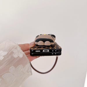 May include: A brown and black phone case with a leopard print design. The case features a 3D cartoon character on top. The case has a brown strap attached to it.