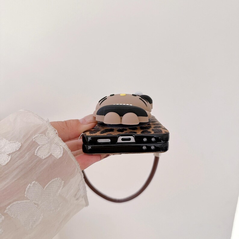 May include: A brown and black phone case with a leopard print design. The case features a 3D cartoon character on top. The case has a brown strap attached to it.