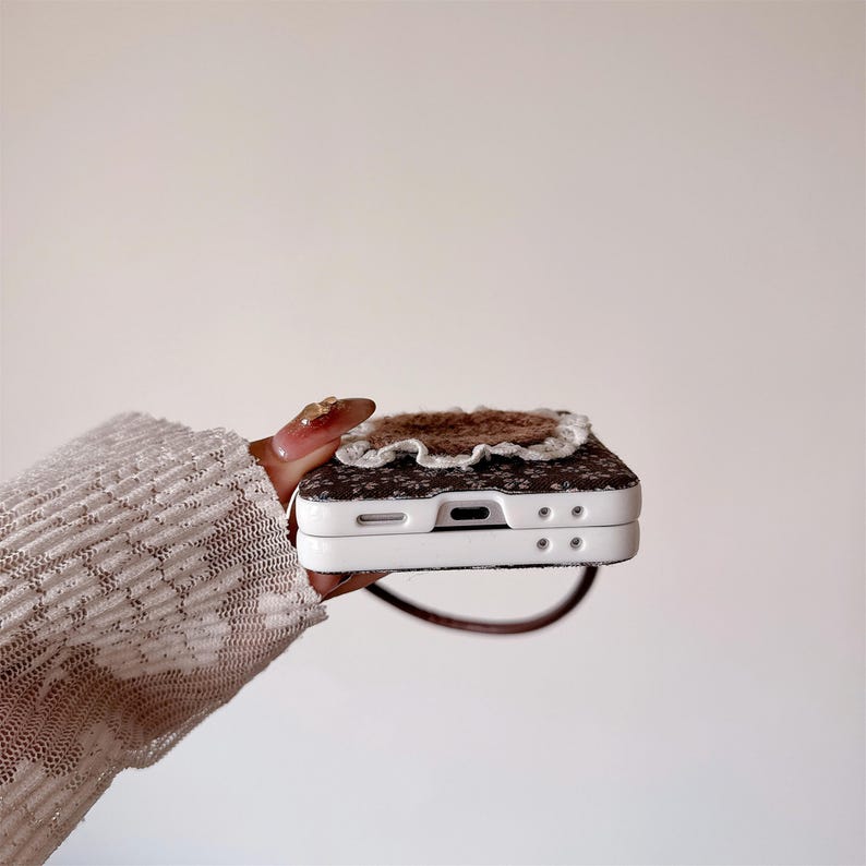 May include: A white and brown floral patterned phone case with a brown and white decorative element on top. The case has a brown strap and is held in a hand.
